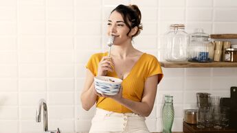 Eine Frau in knallig-gelbem Oberteil sitzt auf der Küchenzeile. Sie hält einen Löffel in der einen und eine Schüssel mit Frühstück in der anderen Hand.