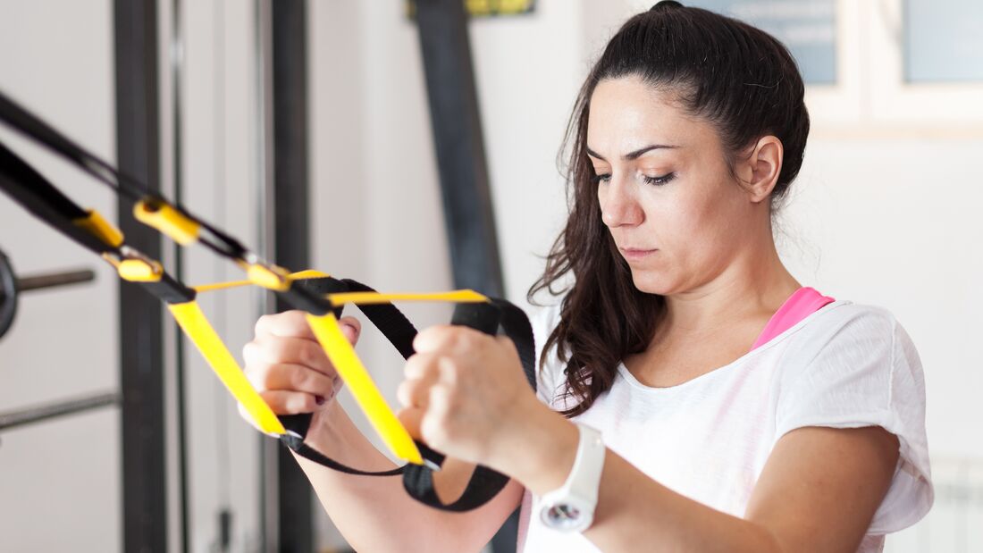 Eine Frau in weißem Shirt und Zopf arbeitet an einem Schlingentrainer. Sie schaut konzentriert.