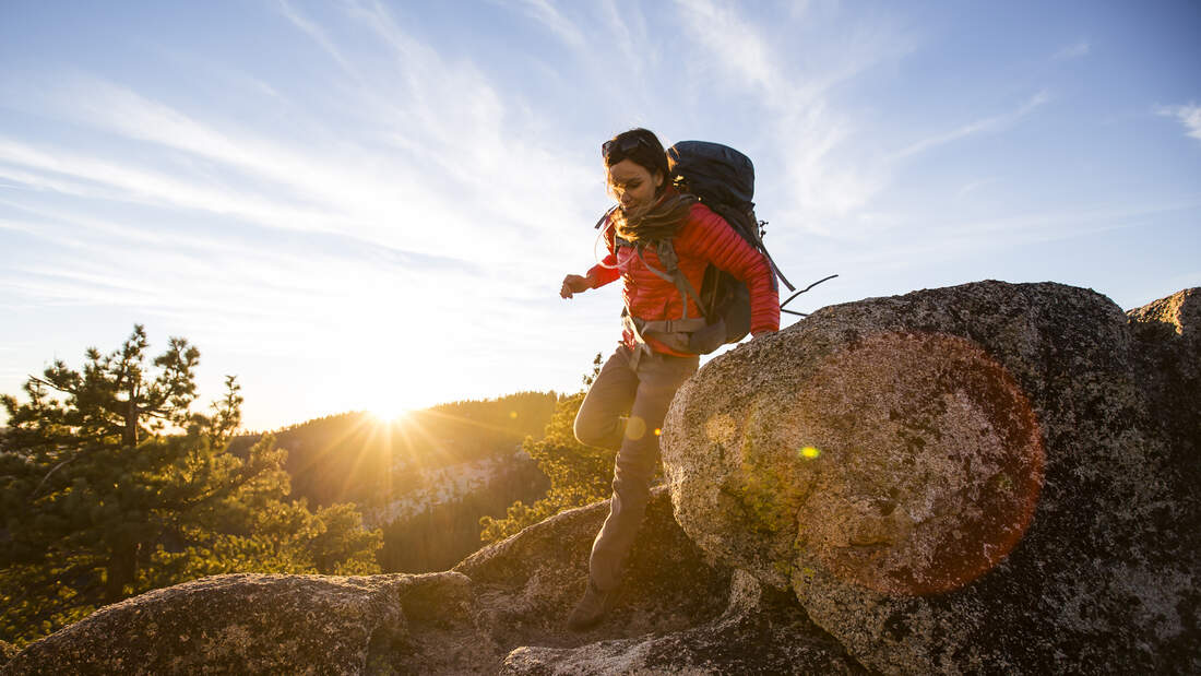 Eine Frau mit Wanderkleidung und großem Rucksack steigt bei Sonnenuntergang einen holprigen Wanderweg hinab.