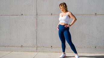 Frau mittleren Alters in Sportbekleidung aus weißem Sport-BH und blauen Leggings vor einer Wand