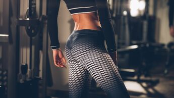 Man sieht den Unterkörper einer Frau in Sportkleidung. Sie steht in einem Gym-Setting, der Fokus liegt auf ihrem Gesäß.