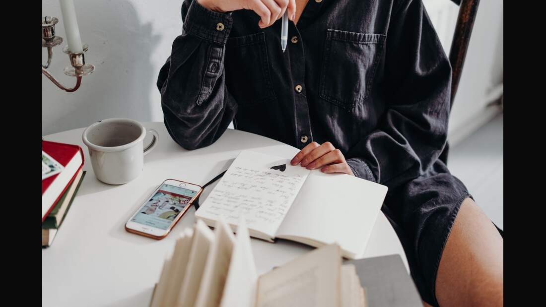 Notizbuch liegt auf Tisch und Frau hält Stift in der Hand.